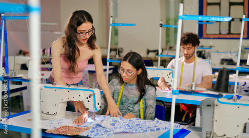 Indian young adult gen z woman lady wear tape around neck talk learn new clothes craft sewing course. Teen pupil girl female sitting desk help to stitch teach casual skill day indoor class room place
