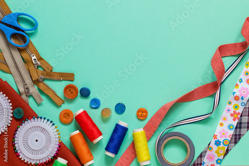 Assorted sewing and tailoring tools arranged on a pastel turquoise background, featuring thread spools, buttons, ribbons, zippers, pins, and scissors. Copy space.