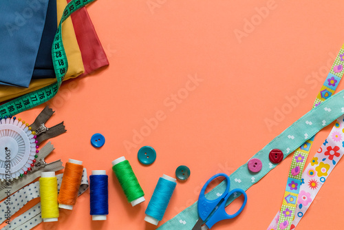 Colorful sewing equipment arranged on a coral surface with thread reels, buttons, zippers, pin wheels, and scissors for dressmaking projects. Copy space.