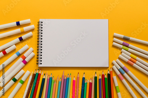 Blank spiral notebook surrounded by colorful markers and pencils on a bright yellow background, creating a creative school and art workspace.