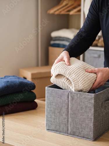Person's hands folding knitted sweaters into fabric storage basket at home. Seasonal wardrobe organization of winter clothes. Household chores, decluttering and tidying concept.