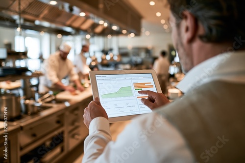 Over-the-shoulder view of a male restaurant manager in white shirt and vest holding a tablet displaying electricity usage charts and efficiency metrics while chefs work at stainless steel preparation