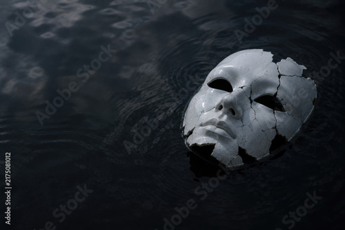 Cracked white face mask floats on dark water. Space for text.