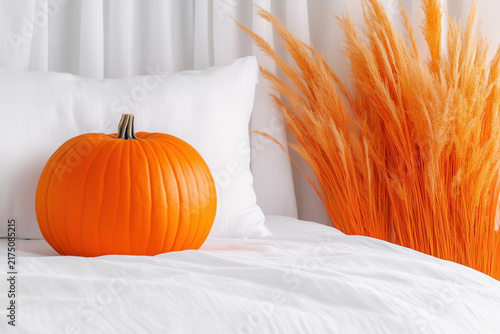 Orange pumpkin with white pillow and pampas grass on bed with copy space.