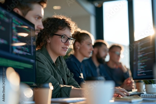 Diverse group of young male and female software developers intensely focused on computer screens displaying colorful lines of code in a modern office workspace with large windows letting in bright