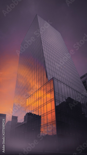 Modern glass skyscraper facade reflecting vibrant orange and purple sunset sky over misty urban landscape at twilight