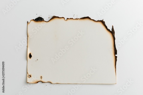 Blank sheet of heavy paper with irregular scorched edges and small charcoal blackened sections resting on seamless white surface, fine ash particles scattered naturally around perimeter while