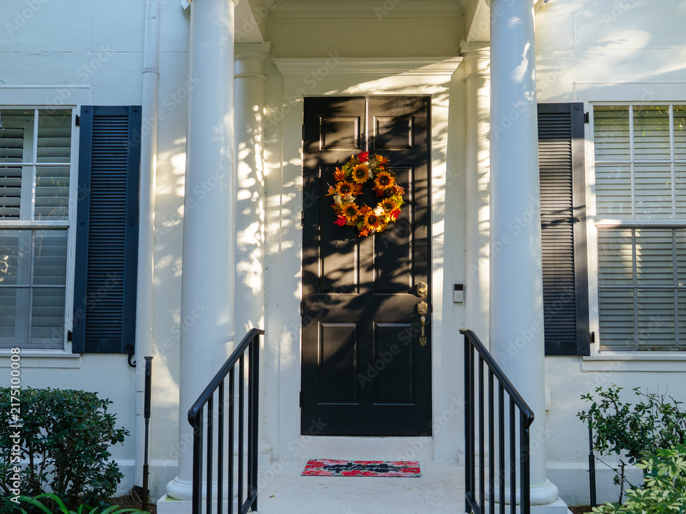 Naklejka premium A welcoming home entrance features a colorful autumn wreath on a black front door framed by elegant white columns. Soft afternoon light creates a warm seasonal atmosphere.