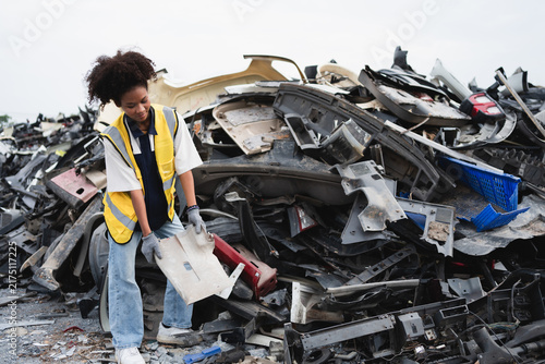 A recycling worker inspects and handles a recovered vehicle component among discarded car parts, representing automotive recycling and resource recovery