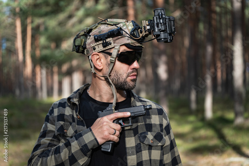 фотографія Focused tactical operator wearing a night vision cap with an attached thermal imager monocular and a separate optical night vision monocular holding a handgun outdoors