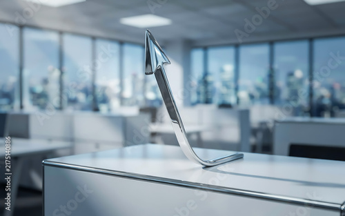 Chrome metallic upward growth arrow object on desk surface in bright modern open plan corporate office space