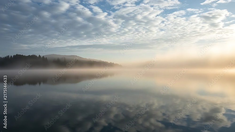 Naklejka premium Morning mist rising over calm lake surface with cloud reflections.