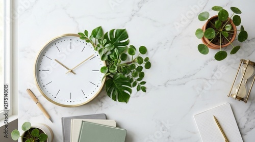 Minimalist white clock decorated with fresh green tropical leaves on a marble desk workspace with notebooks