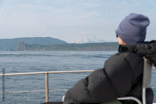 Fotografie Woman in dark jacket and knit hat relaxes on catamaran overlooking Pacific Ocean, Kamchatka coast and distant volcano