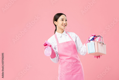 Smiling housekeeper with cleaning supplies on pink background