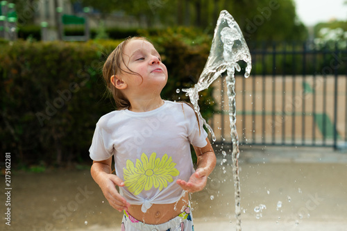 A young girl in a wet white t-shirt plays with a spraying water fountain in Paris, France. Water splashes in the air as she tilts her head back outdoors.