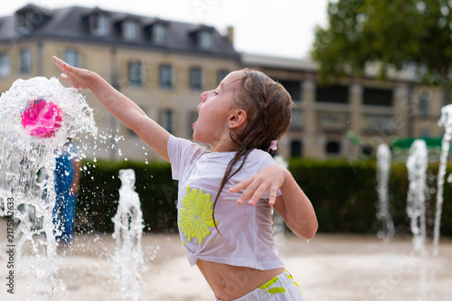 A young girl in a wet white t-shirt plays with a pink ball over a splashing water fountain in Paris, France. She reaches her arm out to touch the water stream outdoors.
