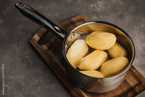 Peeled raw potatoes in a stainless steel saucepan with water on wooden cutting board