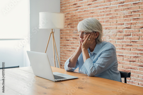 Foto Senior woman rests chin on hands at a wooden table with laptop, staring away wit