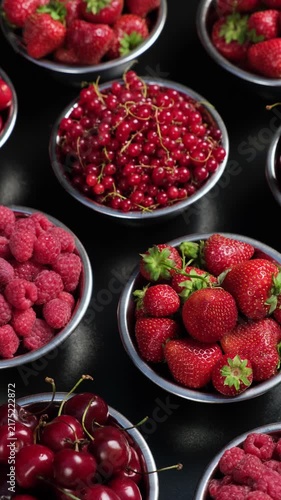Vertical top view of assorted fresh berries including raspberries currants strawberries and cherries in bowls on a dark background rotation