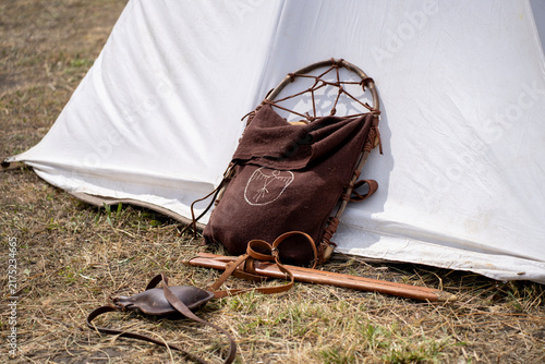 Konstfotografi Authentic historical reenactment equipment including medieval frame backpack, leather sword scabbard, and flask lying near a white canvas camp tent