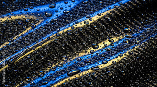 Water droplets on textured carbon fiber with blue and yellow light reflections.
