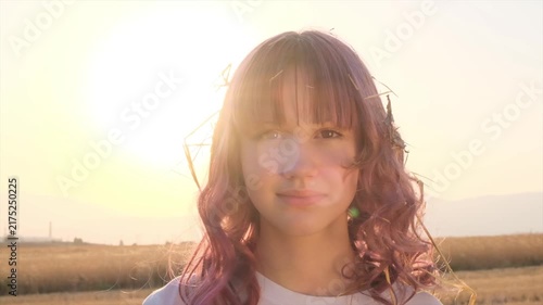 Preteen girl portrait at sunset, smiling with voluminous light brown hair and straw caught in her hairstyle, natural child expression, rural lifestyle, summer countryside, warm evening light