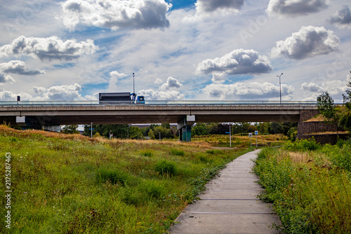 Plain with a pedestrian path, elevated motorway bridge with a truck driving in background, against blue sky and white clouds, wild grass, sunny summer day in South of Limburg, Netherlands