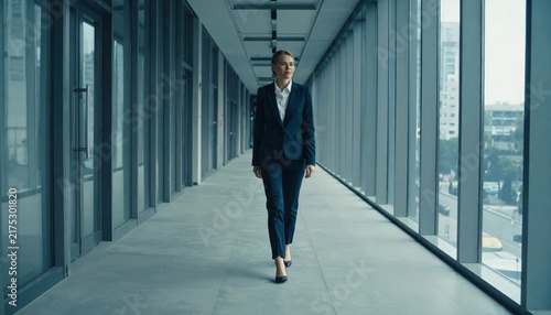 Professional woman walking forward in sleek glass office hallway.