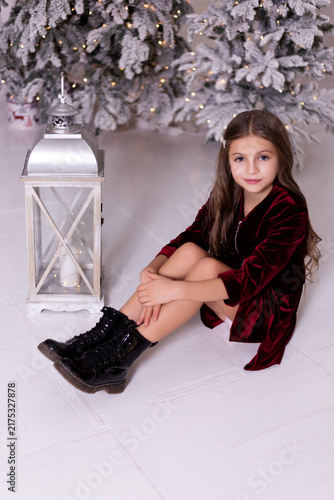 Charming little girl sitting on floor near decorative lantern and snowy Christmas tree