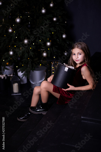 Elegant little girl holding gift box sitting near Christmas tree on dark studio background