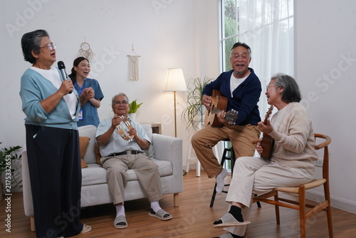 A group of elderly people are preparing for an activity: a senior woman holds a microphone and sings, and her friend plays guitar, enjoying music therapy at the nursing home.