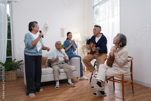 A group of elderly people are preparing for an activity: a senior woman holds a microphone and sings, and her friend plays guitar, enjoying music therapy at the nursing home.
