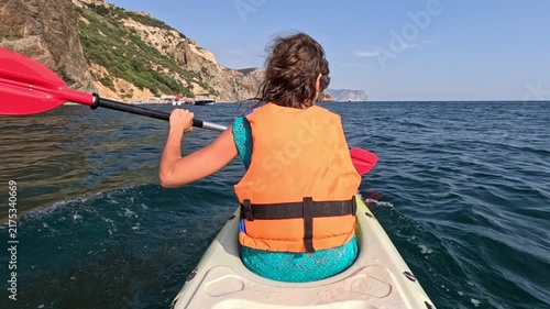 Paddling single kayak along scenic mountainous shore on calm ocean water during sunny afternoon. Rear view tracking shot.
