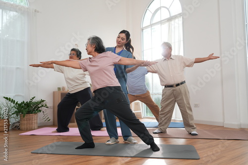 A group of elderly men and women are exercising and practicing yoga, with an Asian female nurse serving as a turner providing physical support and care in a class at the nursing home.