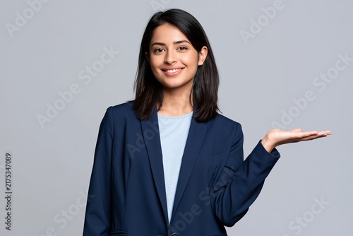 Smiling woman in a blue blazer presenting something with an open hand against a grey wall