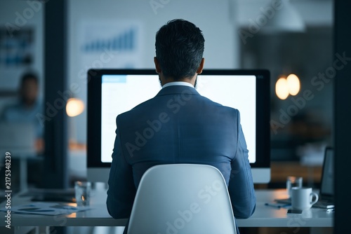 Businessman sitting at desk in front of computer monitor working late in dark office interior