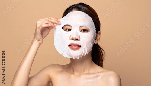 Young woman removing sheet face mask on beige background, skincare concept