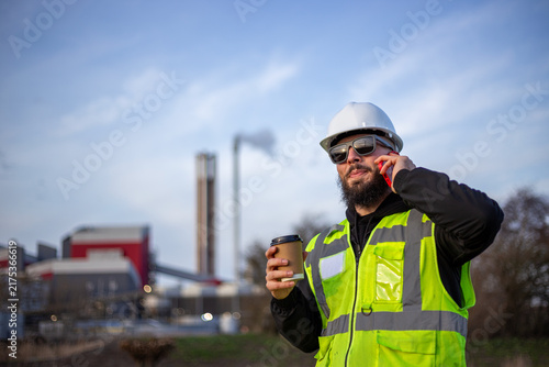 Industrial worker wearing hard hat and safety vest, talking on smartphone while holding coffee cup near power plant