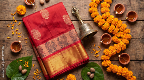 A festive flat-lay of traditional red-bordered saree fabric, brass ghanta, marigold garland, and clay diya
