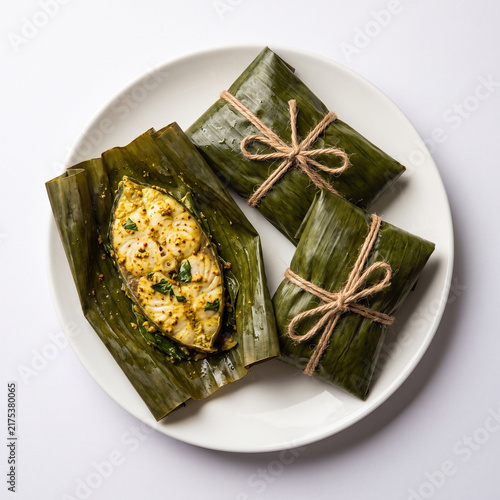 Steamed Bhetki Paturi wrapped in banana leaf tied with thread, one opened slightly showing spiced fish, white background, flat lay