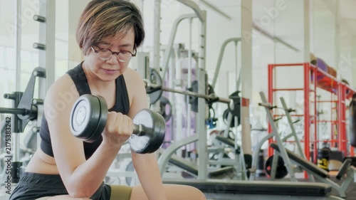 Active Asian woman in her forties wearing glasses and a sports bra lifting dumbbells inside a bright modern gym. She focuses on her mirror reflection during a daily strength training workout.