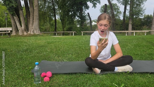 Obraz na płótnie Young girl in a white t‑shirt and black leggings sits cross‑legged on a dark grey yoga mat in a park