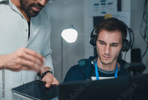 Technology IT Programmer Working At Computer With Headphones While Colleague Uses Tablet In Modern Office, Software Development Team Collaboration, Coding, Technical Support And Digital Business.