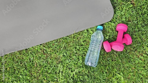 Close‑up outdoor shot showing a dark grey fitness mat partially in frame on the left, its smooth textured surface angled diagonally. On green grass in the foreground — a transparent plastic water bott