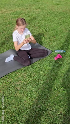 Girl in white t‑shirt and black leggings sitting on grey yoga mat on grass, focused on smartphone. Pink dumbbells and plastic water bottle nearby. Natural daylight, calm outdoor park scene