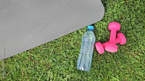 Close‑up outdoor shot showing a dark grey fitness mat partially in frame on the left, its smooth textured surface angled diagonally. On green grass in the foreground — a transparent plastic water bott