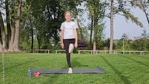 Girl in white t‑shirt and black leggings doing balance exercise on trainer. Pink dumbbells and water bottle on grass. Park with trees, natural daylight