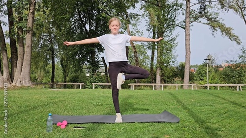 Girl in white t‑shirt and black leggings holding wide balance pose on trainer. Pink dumbbells and water bottle on grass. Park with tall trees, bright daylight