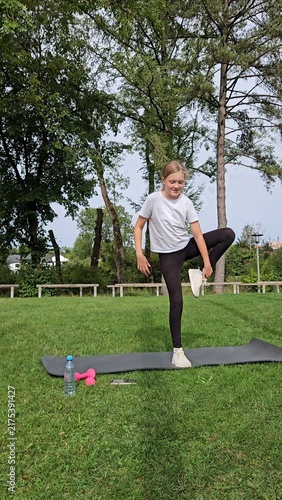 Girl in white t‑shirt and black leggings holding balance pose on trainer. Pink dumbbells and water bottle on grass. Park with trees, bright daylight